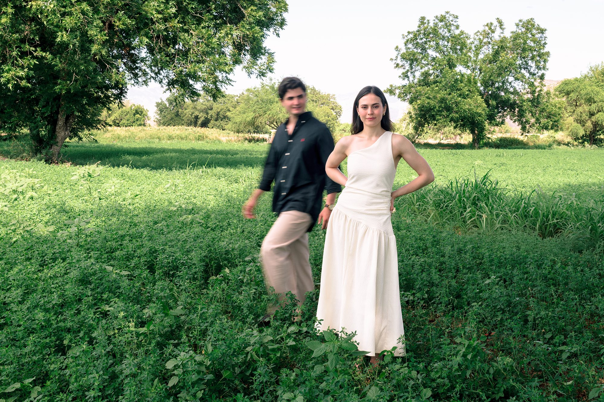 Bride-to-be standing still in a green field while her fiancé walks behind her in motion blur — documentary engagement photography Torreón