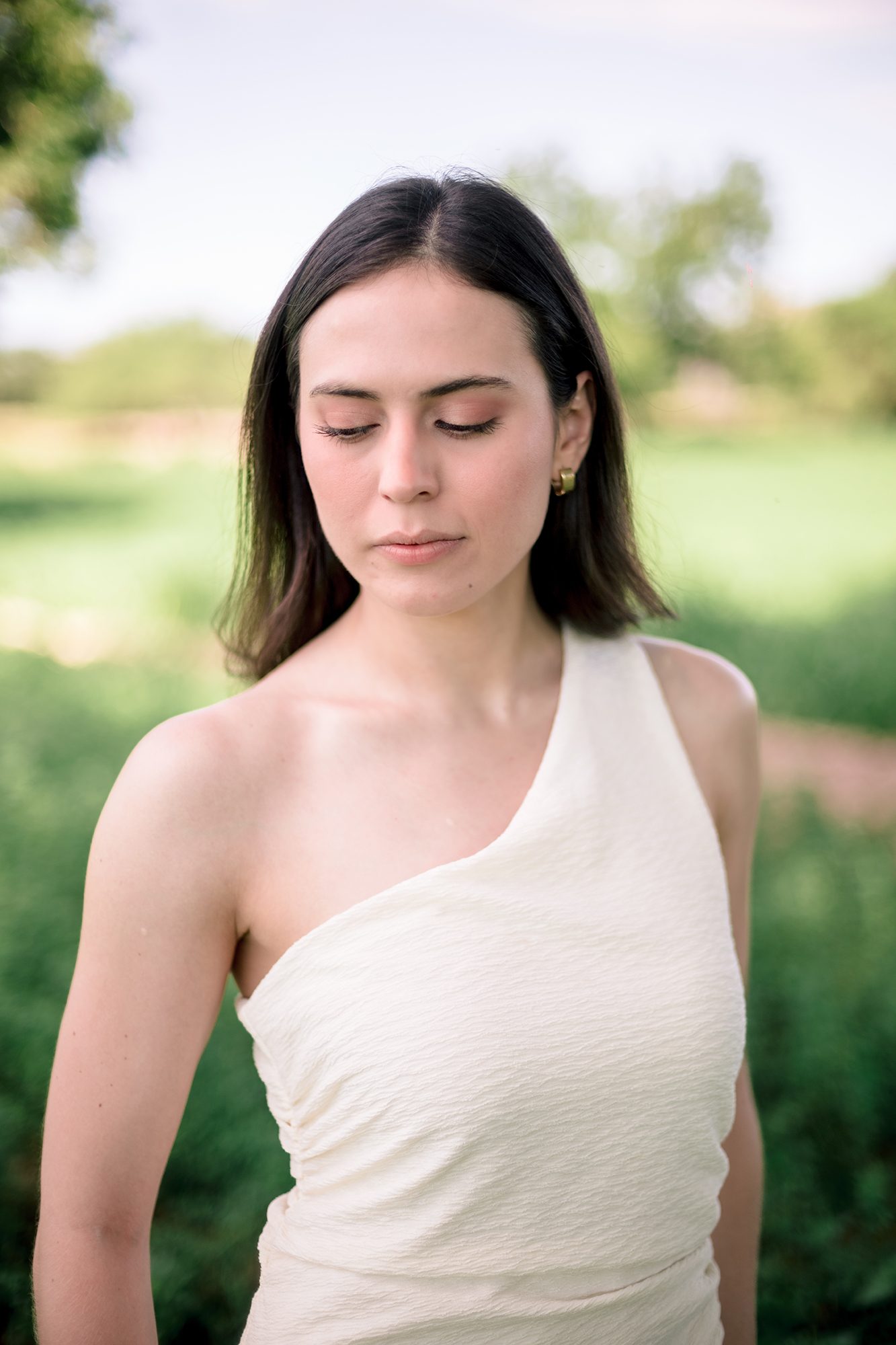 Natural light portrait of a bride-to-be in a white one-shoulder dress, eyes lowered, in a green field — David Lack engagement session Mexico