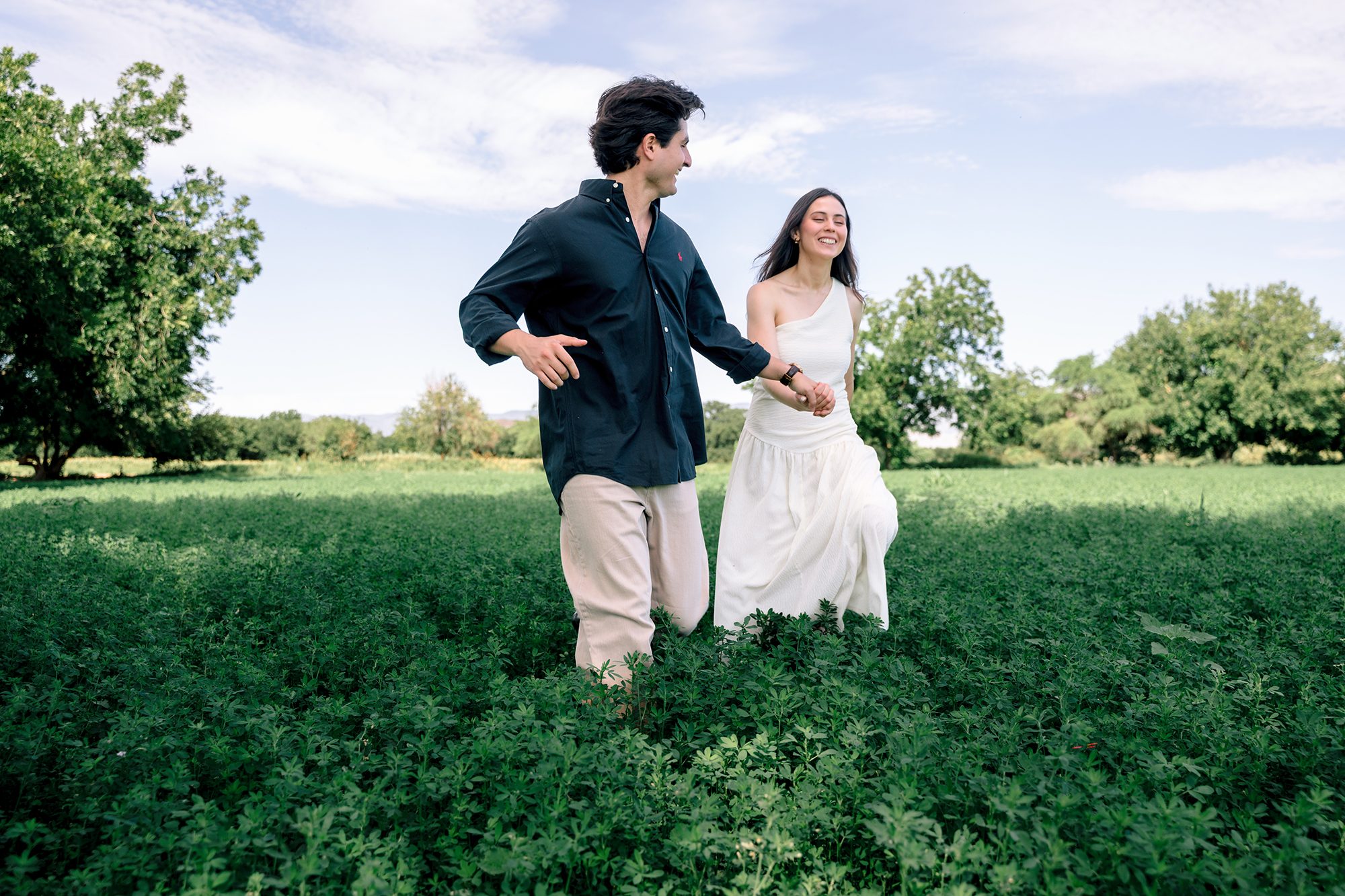 Engaged couple running hand in hand through an alfalfa field in northern Mexico, laughing — David Lack engagement session